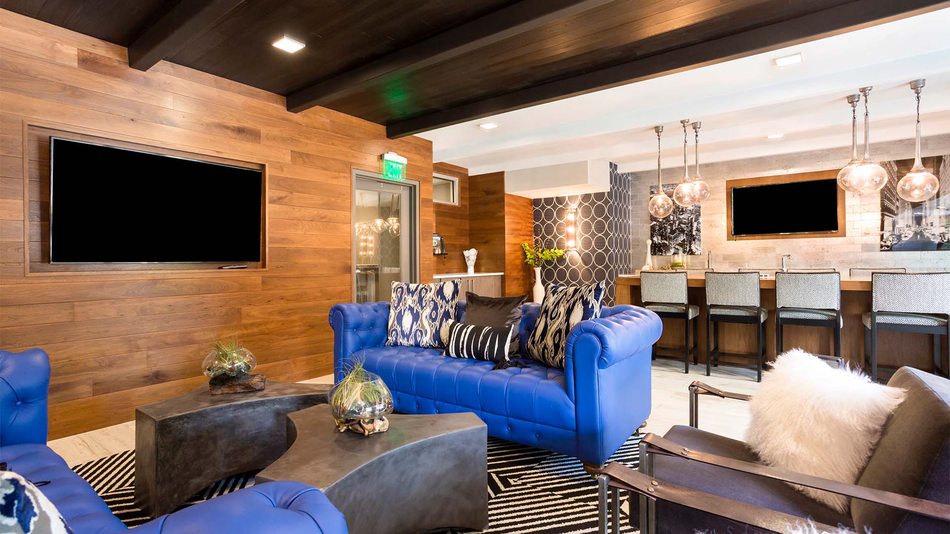 Lounge with blue couches, wood panel wall with mounted TV, breakfast bar with stools, and eclectic decor.