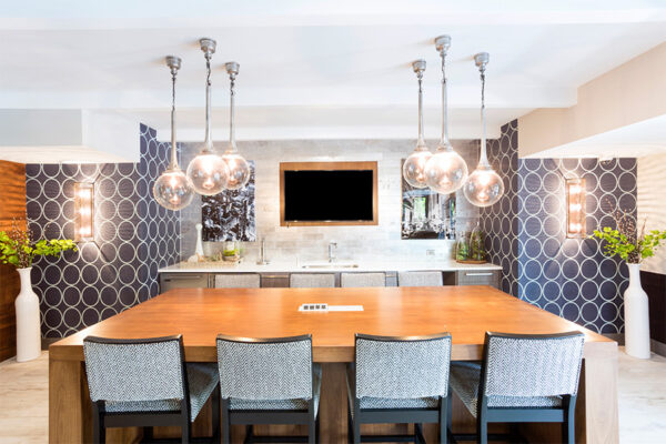 Community kitchen with large dining table, stainless steel appliances, wall mounted TV, and round pendant lamps.