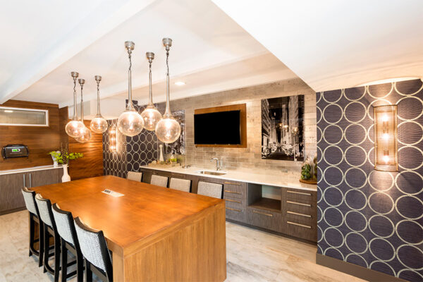 Community kitchen with large dining table, stainless steel appliances, wall mounted TV, and round pendant lamps.