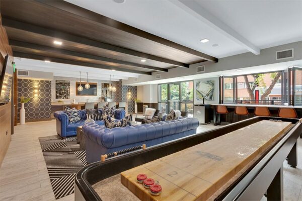 Clubroom with wood floor, plush leather couches, shuffleboard table, and bar with windows to patio.