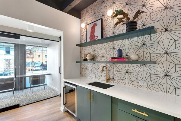 Community kitchen with green cabinets, white counter, bronze hardware, and geometric wallpaper.