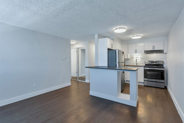 Living area with wood floor and kitchen with white cabinets and stainless steel appliances.
