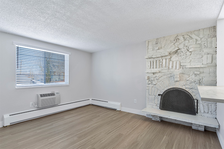 Living room with wood floor, baseboard heaters, white brick accent wall with fireplace, and large windows.