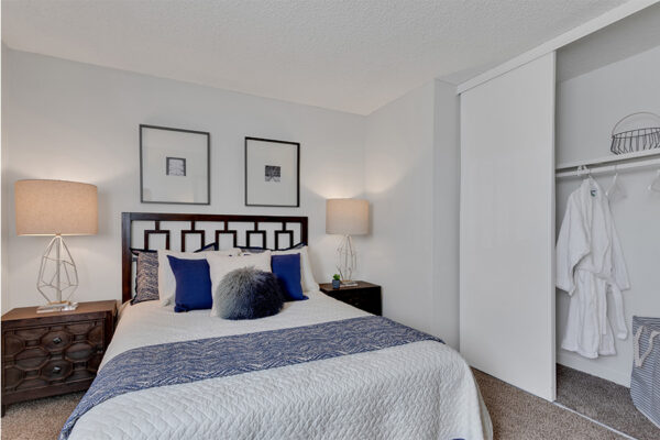 Apartment bedroom with carpet, plush bed, wood bedside cabinets, and wall art.