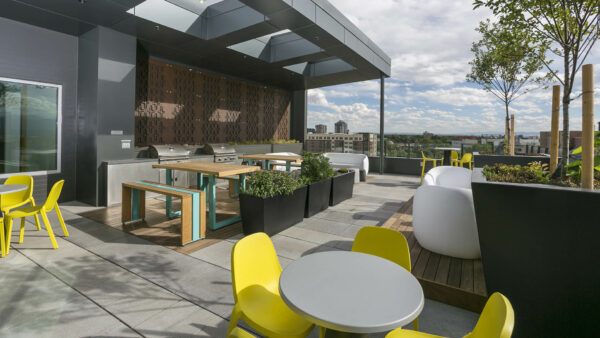 Rooftop patio with outdoor furniture, potted trees, built in BBQ grills, and tall picnic tables.