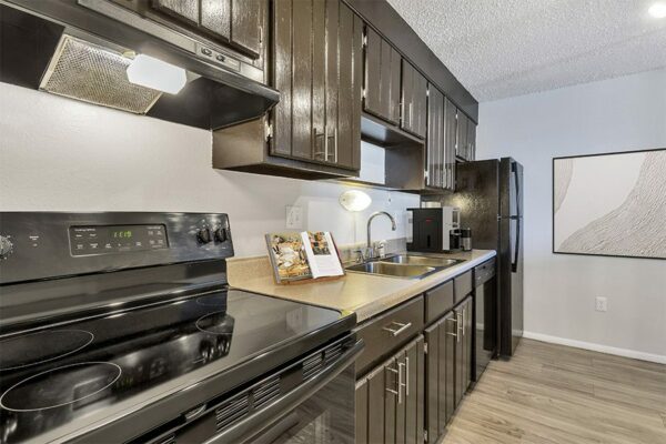 Apartment kitchen with wood floor, brown cabinets, tan counters, and black appliances.