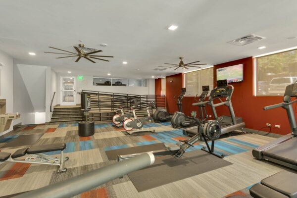 Fitness center with colorful carpet, ceiling fans, wall mounted TV, and cardio machines.