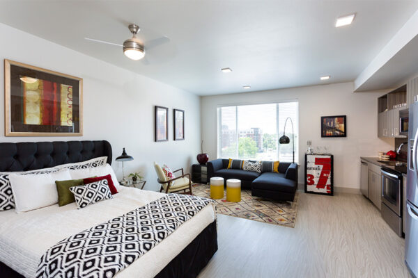Studio apartment with kitchen, plush sectional, large bed, and bright windows.