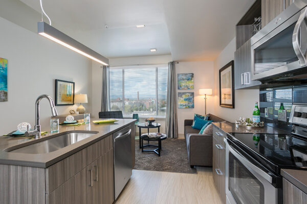 Apartment kitchen and living space with wood floor and cabinets, stainless steel appliances, modern furniture, and large window.