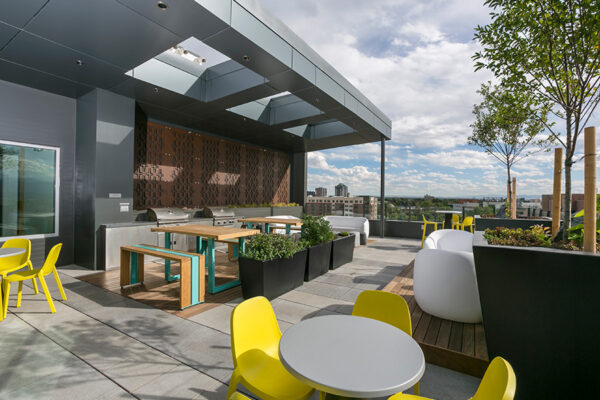 Rooftop patio with outdoor furniture, potted trees, built in BBQ grills, and tall picnic tables.