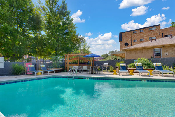 Pool area with bright blue water and lounge chairs under tall trees and cloudy blue skies.