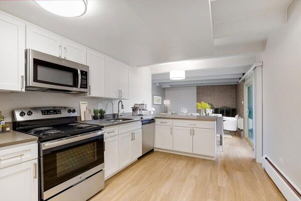 Kitchen with wood floor, white cabinets, grey counters, stainless steel appliances, and living space beyond.