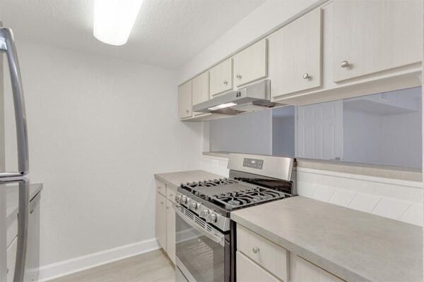 Kitchen with wood floor and cabinets, tan counters, stainless steel appliances, and opening to living area.