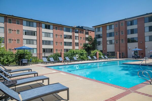 Fenced pool area with lounge chairs, tables with umbrellas, and tall trees overlooked by apartments.