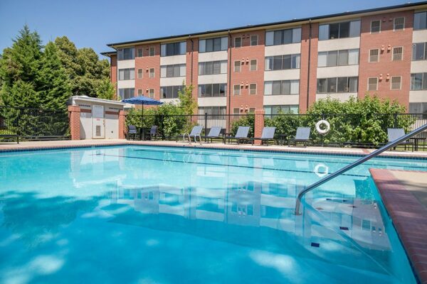 Fenced pool area with lounge chairs, tables with umbrellas, and tall trees.