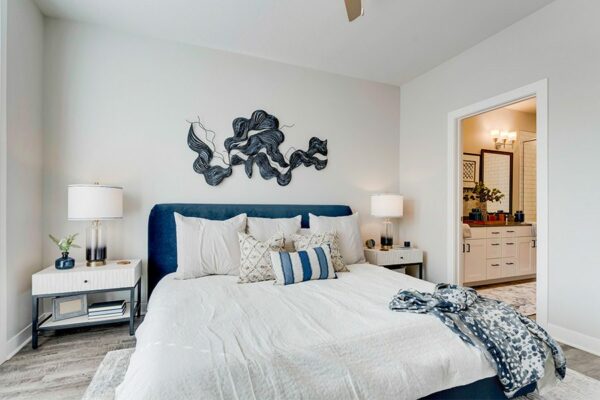 Bedroom with large bed with plush headboard, bedside tables with lamps, flowing blue artwork, and door to bathroom.