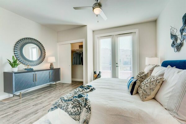 Bedroom with wood floor, large bed, modern furniture, round decorative mirror, and doors to closet and balcony.