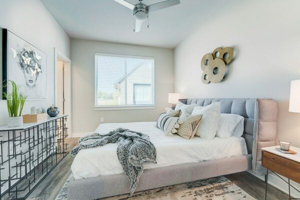 Bedroom with large bed with plush headboard, modern furniture and artwork, ceiling fan, and large window.