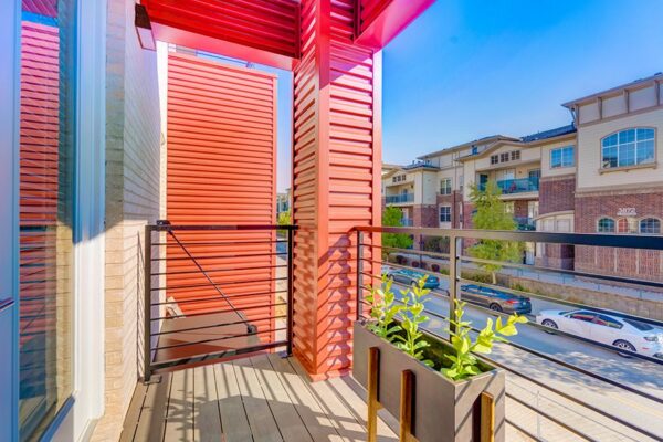 Balcony with potted plant, metal railings, and red metal panels overlooking street parking.