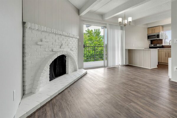 Living room with wood floor, white brick fireplace, kitchen, and door to balcony.