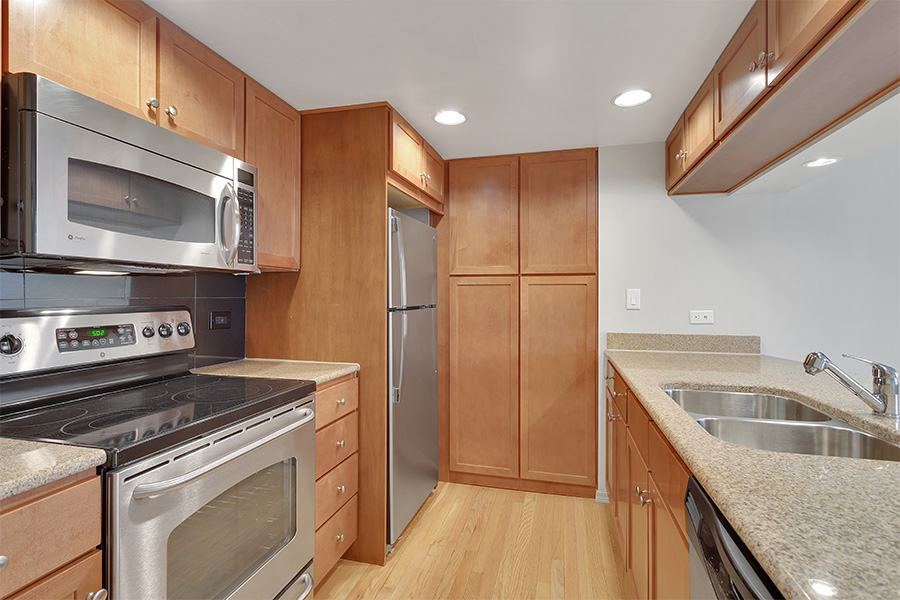 Apartment kitchen with wood floor and cabinets, stone counters, and stainless steel appliances.