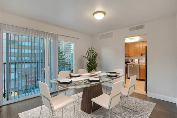 Dining room with wood floor, modern glass table with chairs, potted plant, and doors to balcony and kitchen.