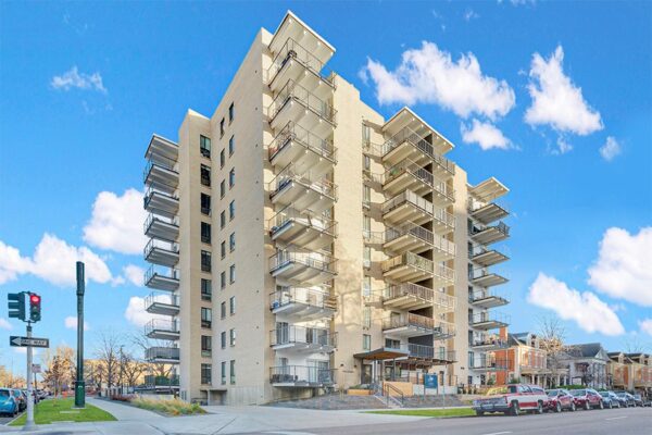 Story apartments with blonde brick construction, large balconies, and garage entrance under cloudy blue skies.