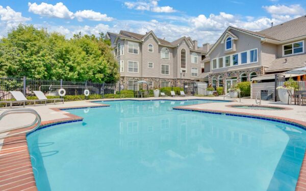 Pool area with brick edging, lounge chairs, tables, and umbrellas overlooked by apartments.