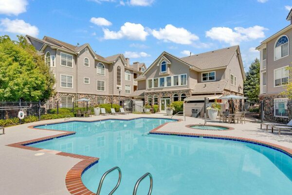 Pool area with brick edging, lounge chairs, tables, and umbrellas overlooked by apartments.
