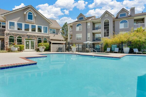Pool area with brick edging, lounge chairs, tables, and umbrellas overlooked by apartments.