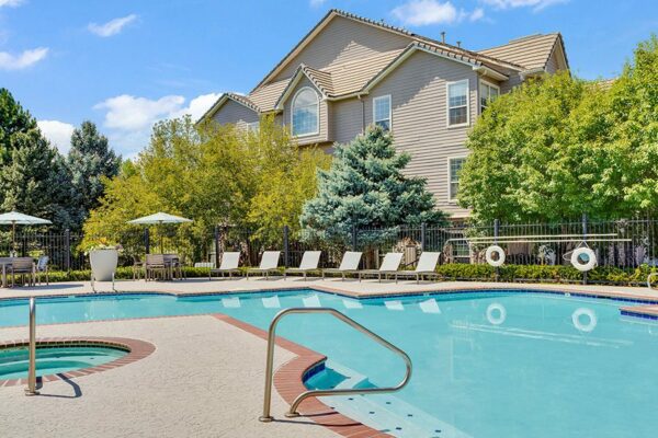 Pool area with brick edging, lounge chairs, tables, and umbrellas overlooked by apartments.