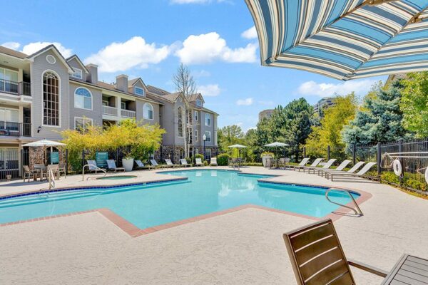 Pool area with brick edging, lounge chairs, tables, and umbrellas overlooked by apartments.