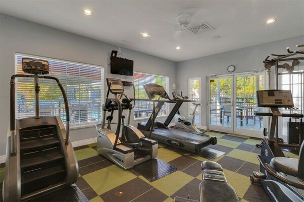 Fitness center with wall mounted TV, cardio machines, weight equipment, and large windows.