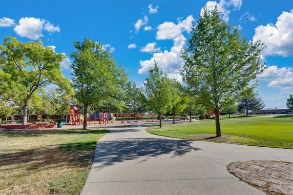 Park with play equipment, lush grass, winding sidewalks and tall trees under cloudy blue skies.