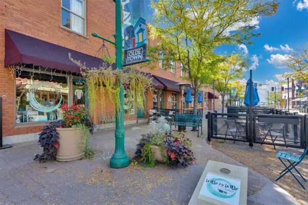 Olde Town Arvada street with stone sidewalk, cornhole game, large potted plants, and tall trees.