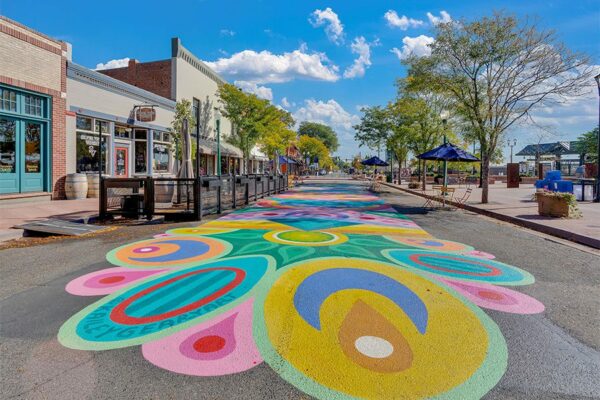 Olde Town Arvada street with sidewalk art, dining patios, and tall trees.