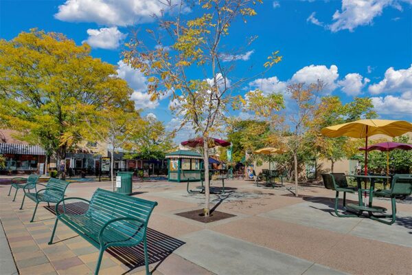 Olde Town Arvada square with benches and outdoor tables under tall trees.