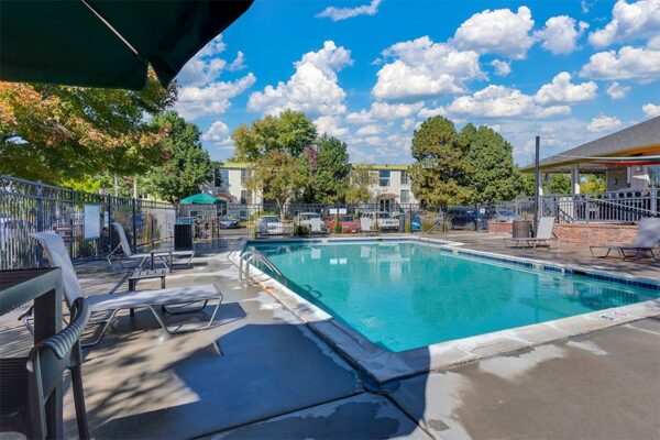 Fenced pool area near clubhouse with lounge chairs and tall trees.