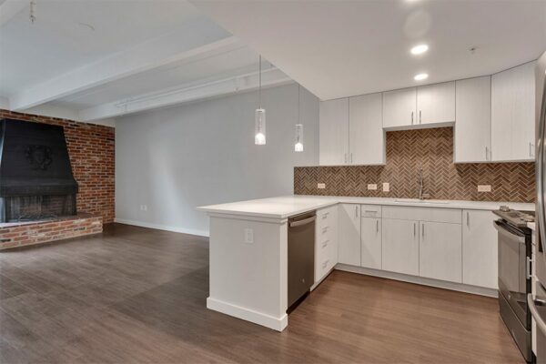 Living area with wood floor, modern kitchen with breakfast bar, and living room with brick accent wall and fireplace.