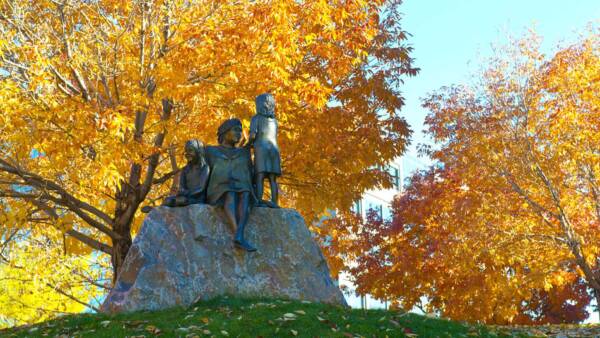 Bronze sculpture of children on rock in grassy field surrounded by tall autumnal trees.