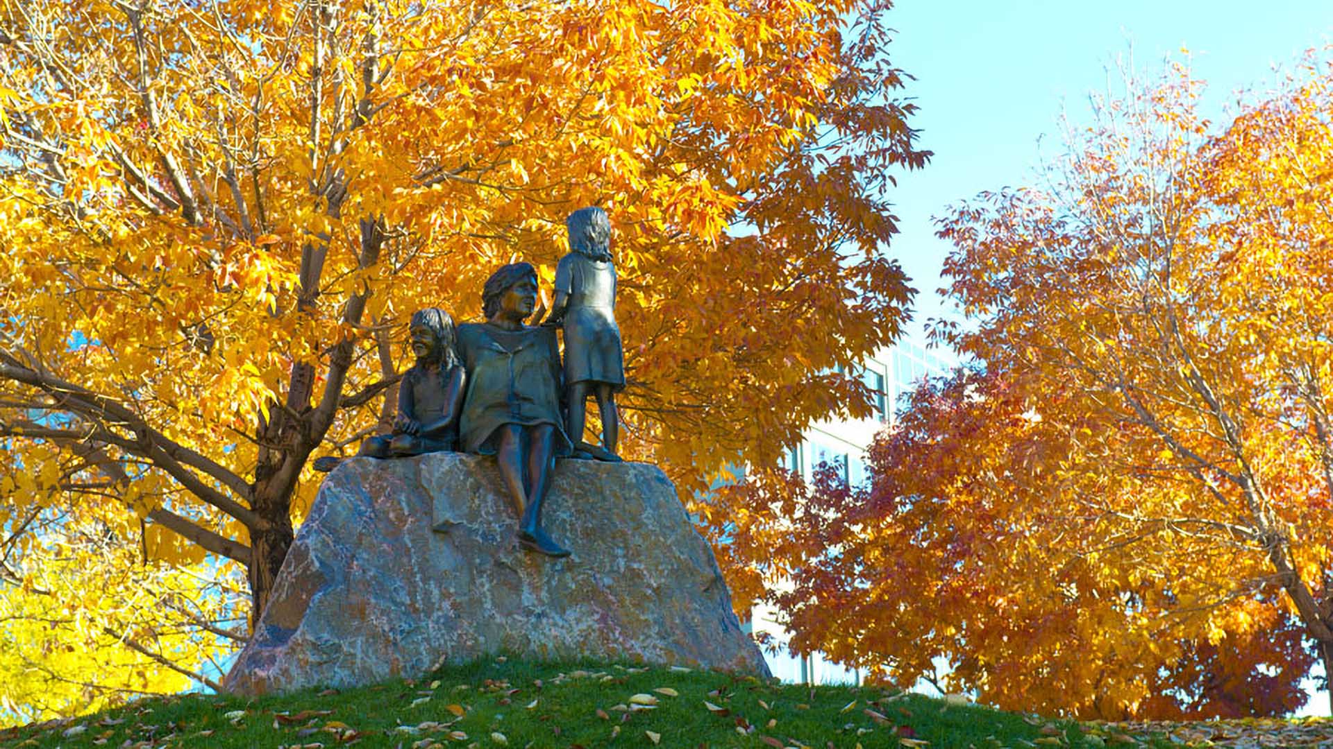 Bronze sculpture of children on rock in grassy field surrounded by tall autumnal trees.