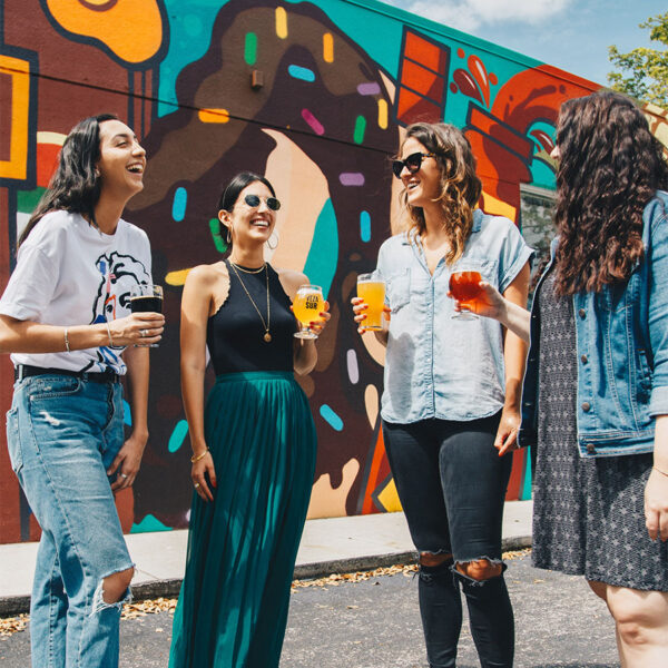 Friends laughing and drinking beer in front of colorful wall mural.
