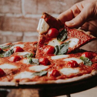 Person picking up slice of thin crust wood fired pizza with tomatoes, mozzarella, and basil.