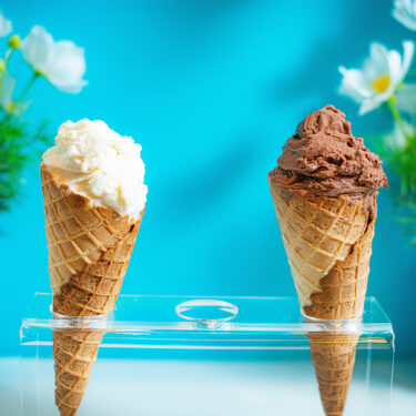Chocolate and vanilla ice cream cones in clear plastic holder in front of bright blue wall.