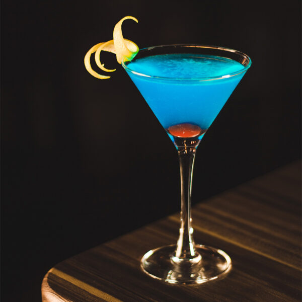 Blue mixed drink in tall martini glass with twist of lemon on dark wood table.