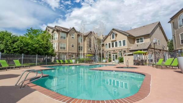 Pool area with brick edging, lounge chairs, tables, and umbrellas overlooked by apartments.