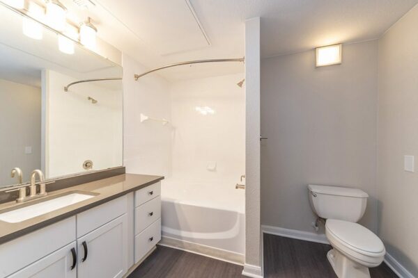 Bathroom with wood floor, white cabinets, grey counter, large mirror, and tiled shower tub.