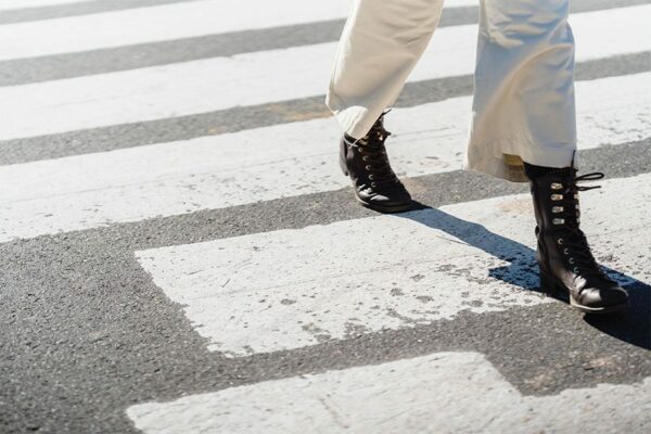 Person in white pants and tall leather boots walking across striped crosswalk.