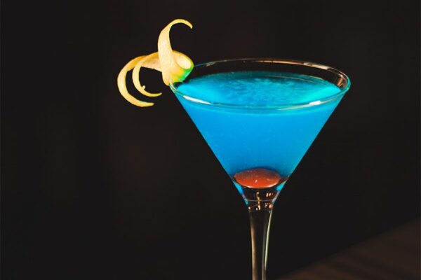 Blue mixed drink in tall martini glass with twist of lemon on dark wood table.
