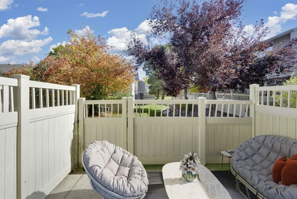 Patio with a light yellow fence by trees and plush chairs.